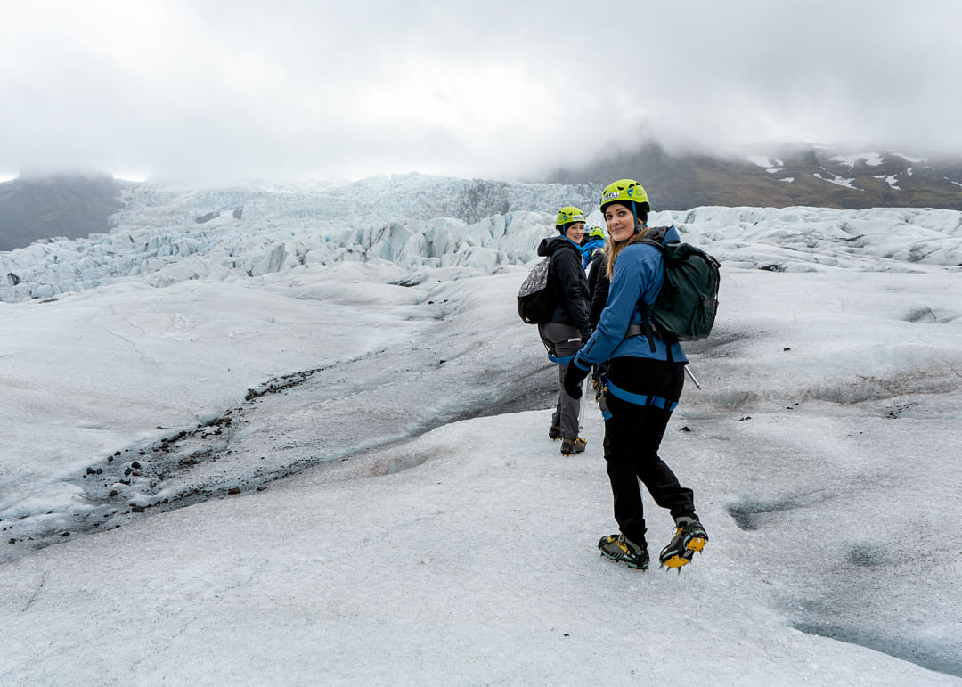 People hiking on a glacier with helmets and gear, surrounded by ice and mountains under a cloudy sky.
