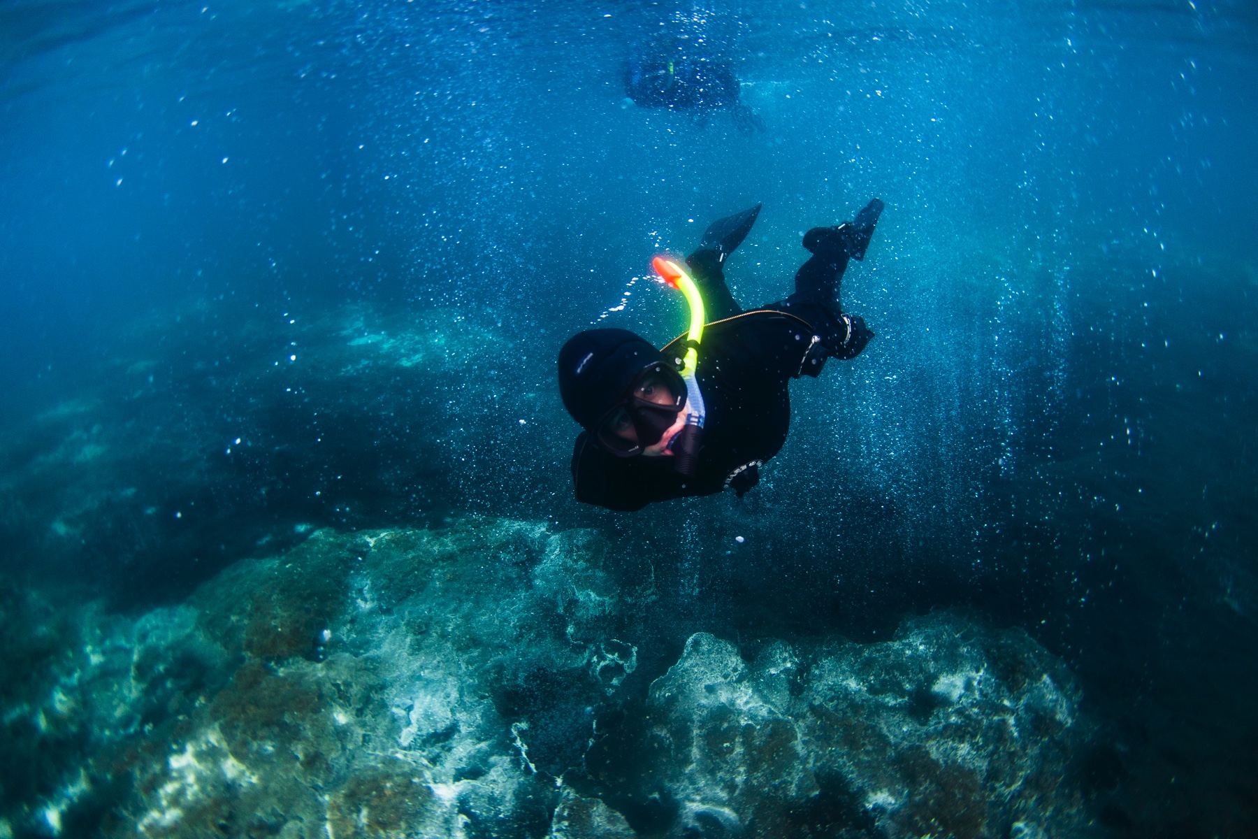 Diver explores underwater depths with snorkel gear, surrounded by clear blue water and rocky ocean floor.