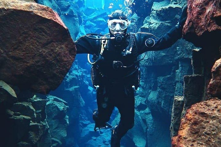 Diver exploring underwater rocks in clear blue water, wearing scuba gear and surrounded by rocky formations.