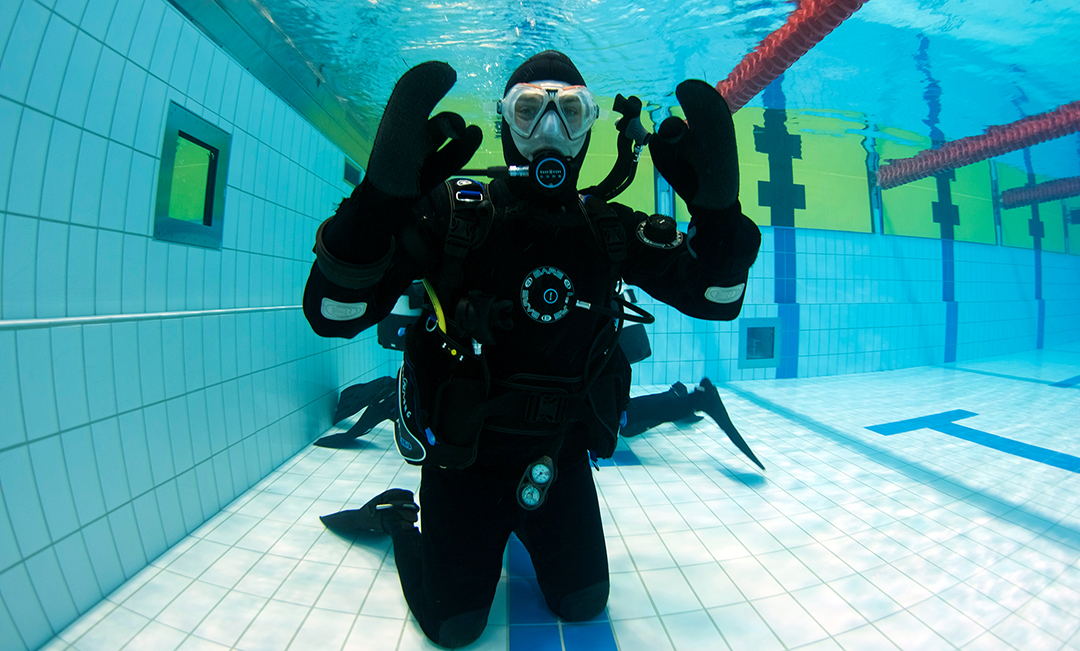Diver practicing scuba skills in an indoor pool, wearing full gear and signaling underwater.