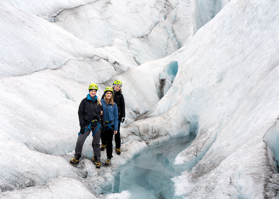 Three people in safety gear standing on an icy glacier, exploring a crevasse with blue ice and water.