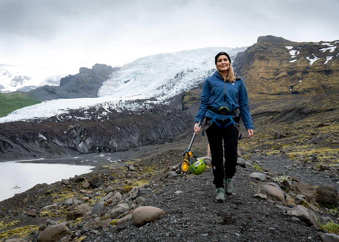Woman in outdoor gear hiking near glacier, carrying climbing helmet and rope on a rocky path.