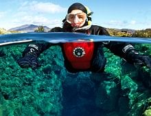 Snorkeler swims in clear blue waters, wearing a red and black wetsuit in a scenic outdoor landscape.