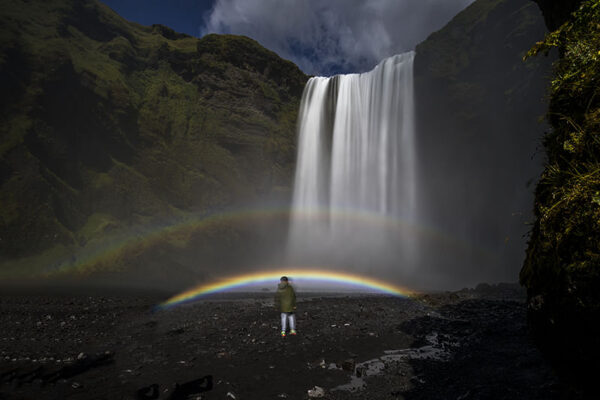 Person standing by a waterfall with a vibrant double rainbow overhead, surrounded by lush greenery.