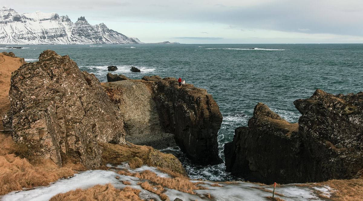 Rugged coastline view with ocean, rocky cliffs, and snowy mountains in the background under cloudy skies.