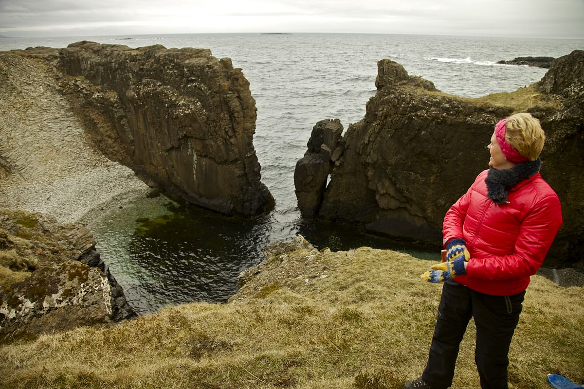 Person in red jacket stands by dramatic coastal cliffs, overlooking ocean waves in overcast weather.