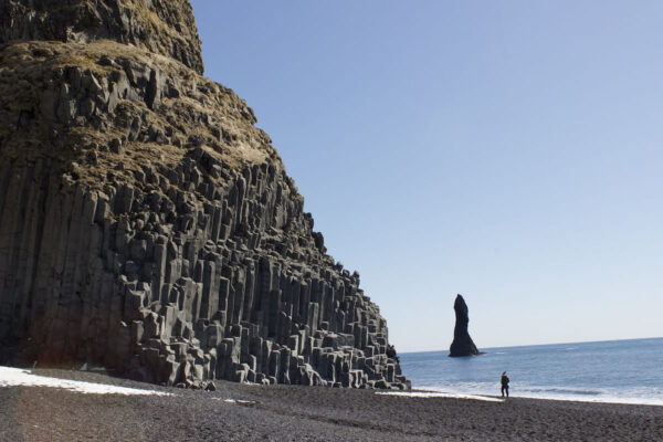 Dramatic basalt columns and rock stack at Reynisfjara Beach, Iceland with a lone figure on the black sand shore.