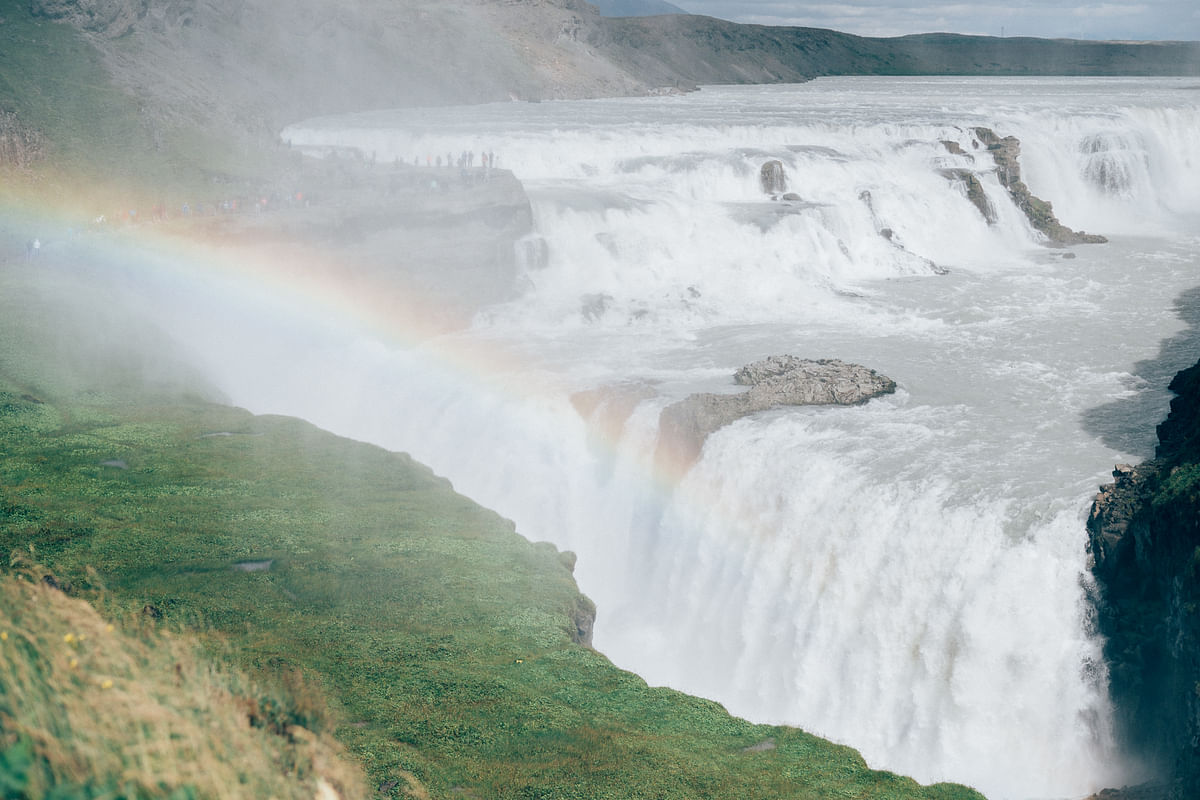 Powerful waterfall cascading into gorge with vibrant rainbow, lush greenery, and misty atmosphere.