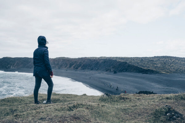Person in a coat overlooking a scenic black sand beach and rocky coastline under a cloudy sky.