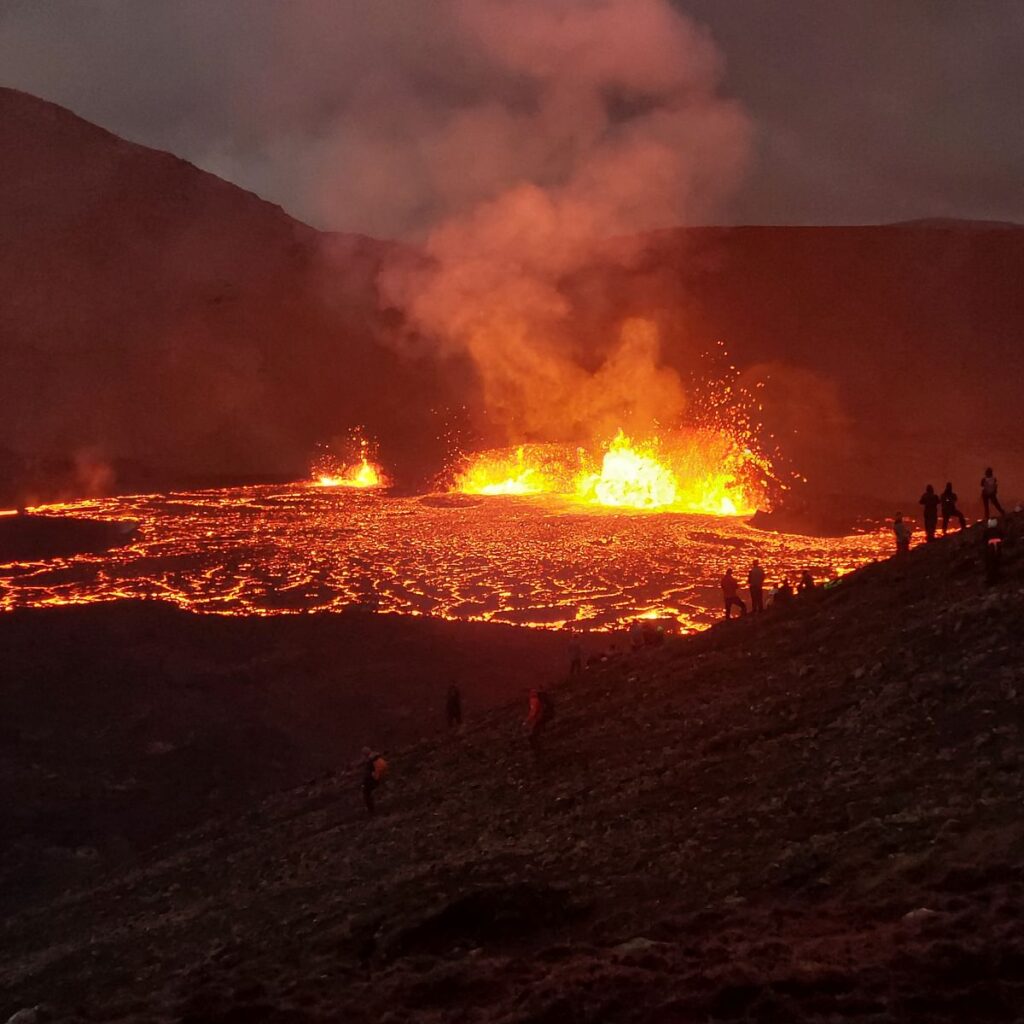 Volcanic eruption with flowing lava and spectators on a hillside at dusk.
