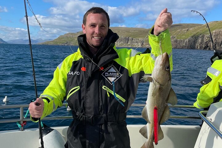 Man on boat proudly holds freshly caught fish against scenic ocean backdrop.