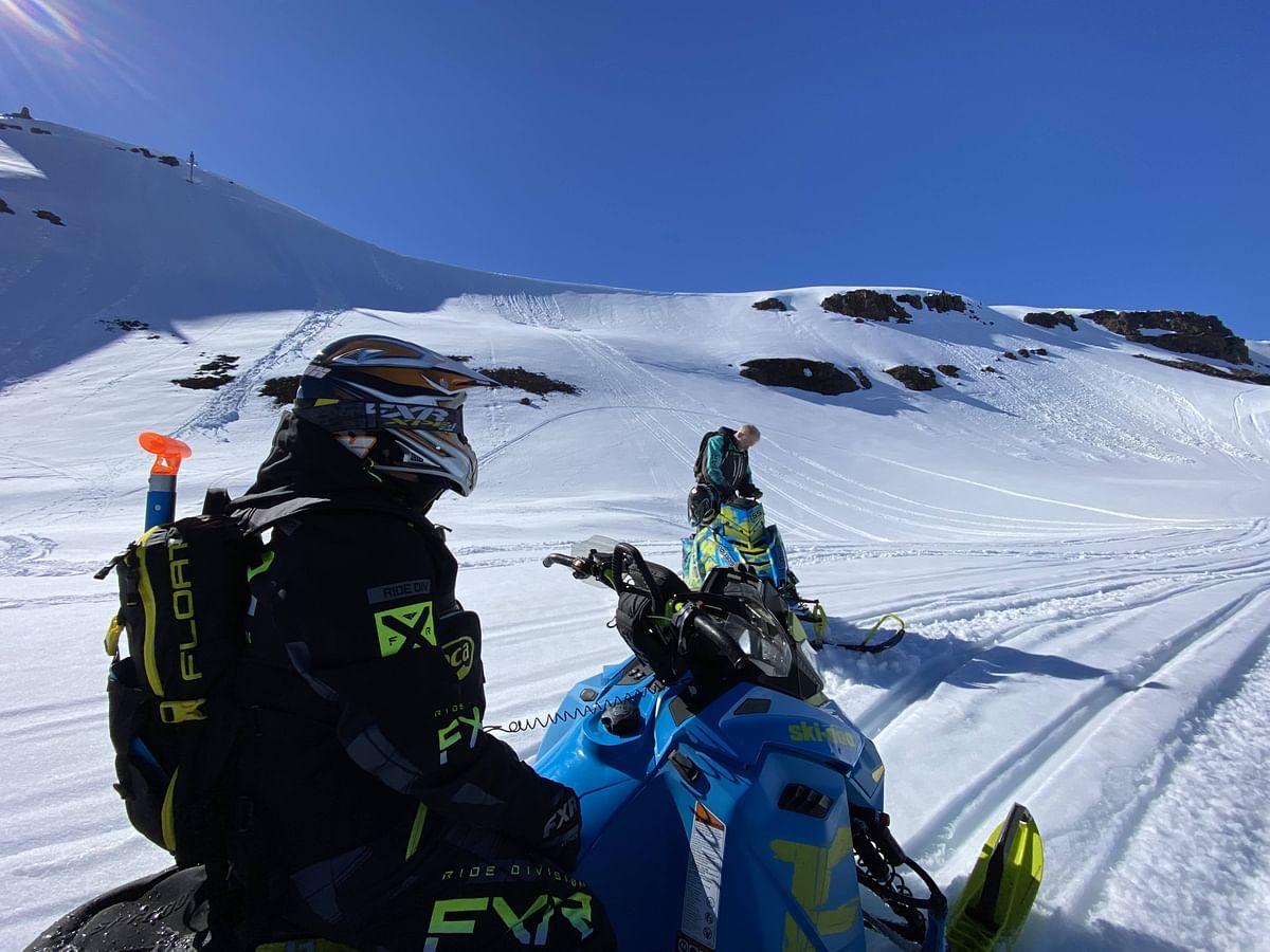 Snowmobilers on snowy mountain terrain under clear blue sky, adventure and winter sports scene.
