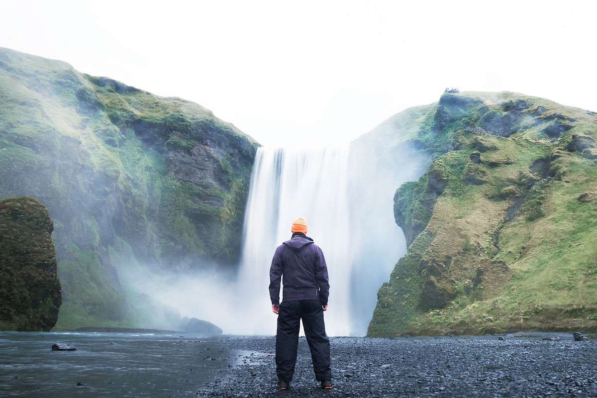 Person in outdoor gear stands before a majestic waterfall surrounded by lush green cliffs.