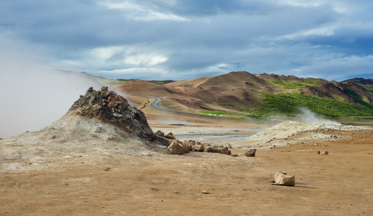 Steaming geothermal landscape with rocky mound, distant road, and grassy hills under cloudy sky.