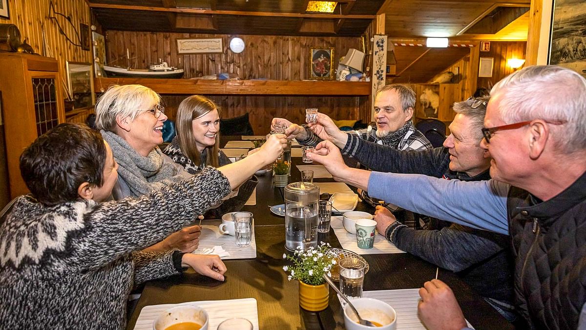 Group of friends toasting with drinks around a cozy dining table in a warm wooden room.