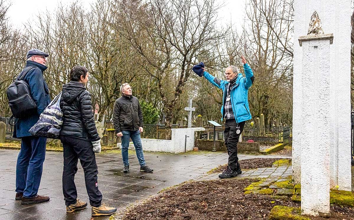 Tour guide gestures to group in a historic cemetery, surrounded by bare trees and graves on a cloudy day.