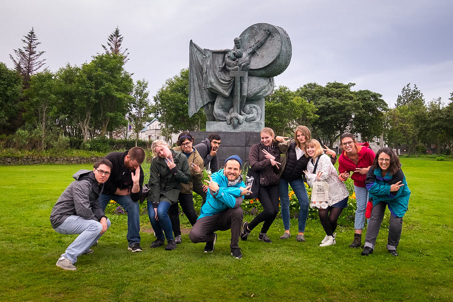 Group posing playfully in front of a large sculpture in a park, with lush greenery and overcast sky in the background.