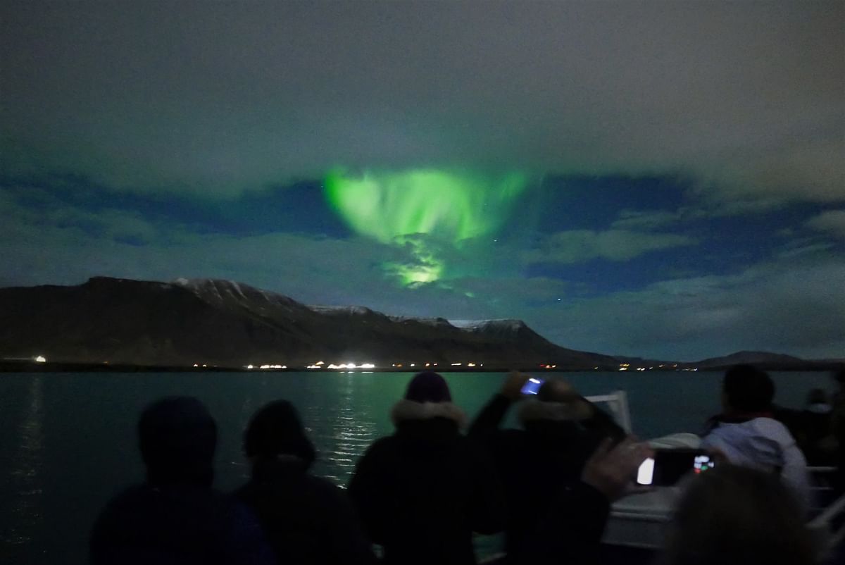 People watch vibrant green aurora borealis dancing over mountains and water under a cloudy night sky.