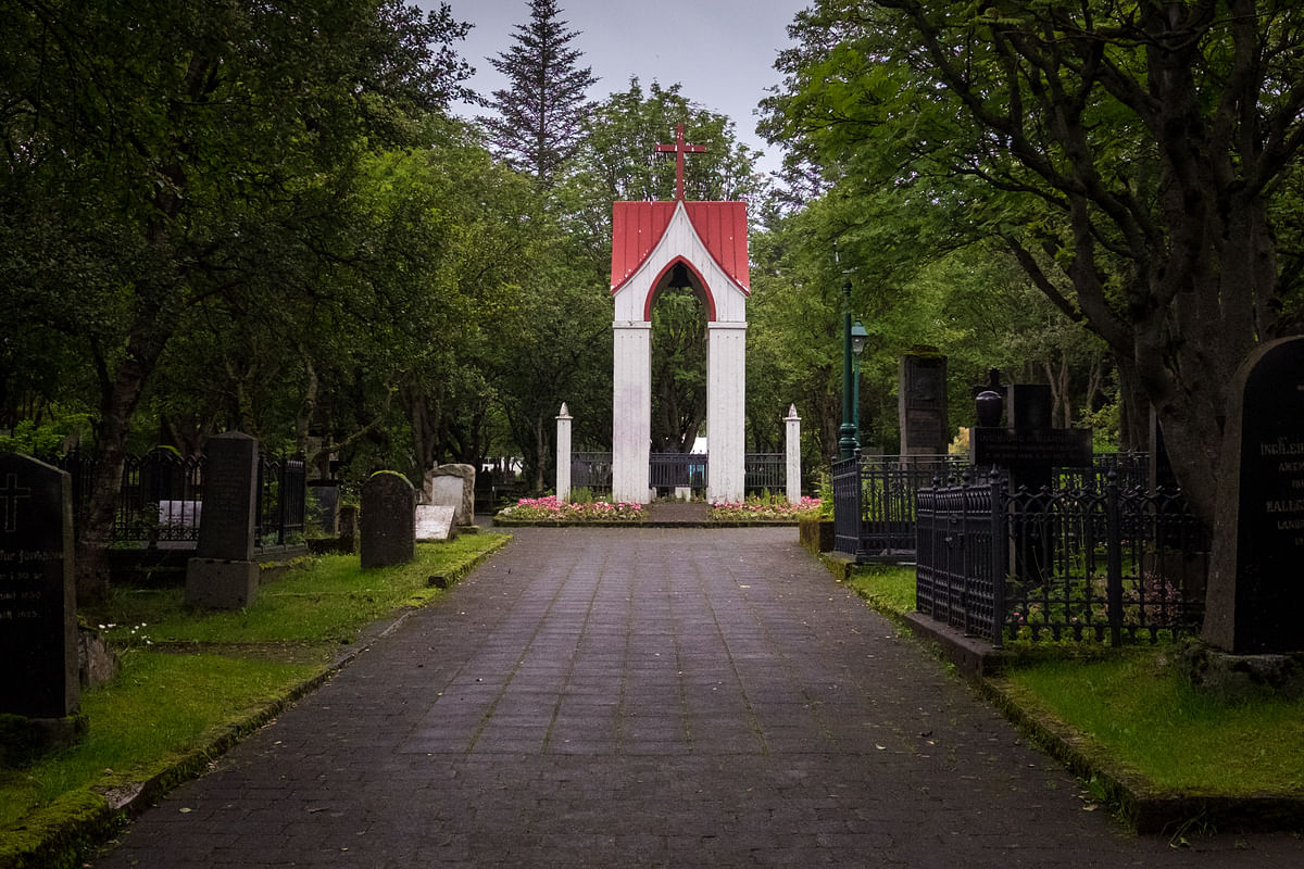 Peaceful cemetery path lined with tombstones, leading to a white arch with a red roof amidst lush greenery.
