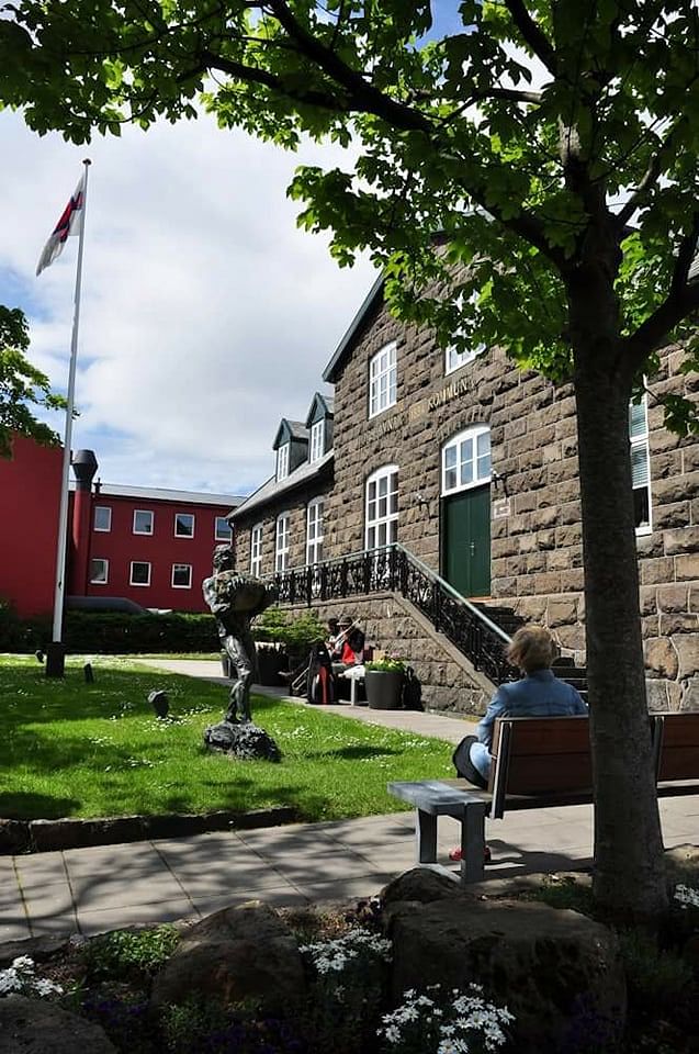 Historic stone building with a garden and statue under a tree, with a person sitting on a bench in sunlight.
