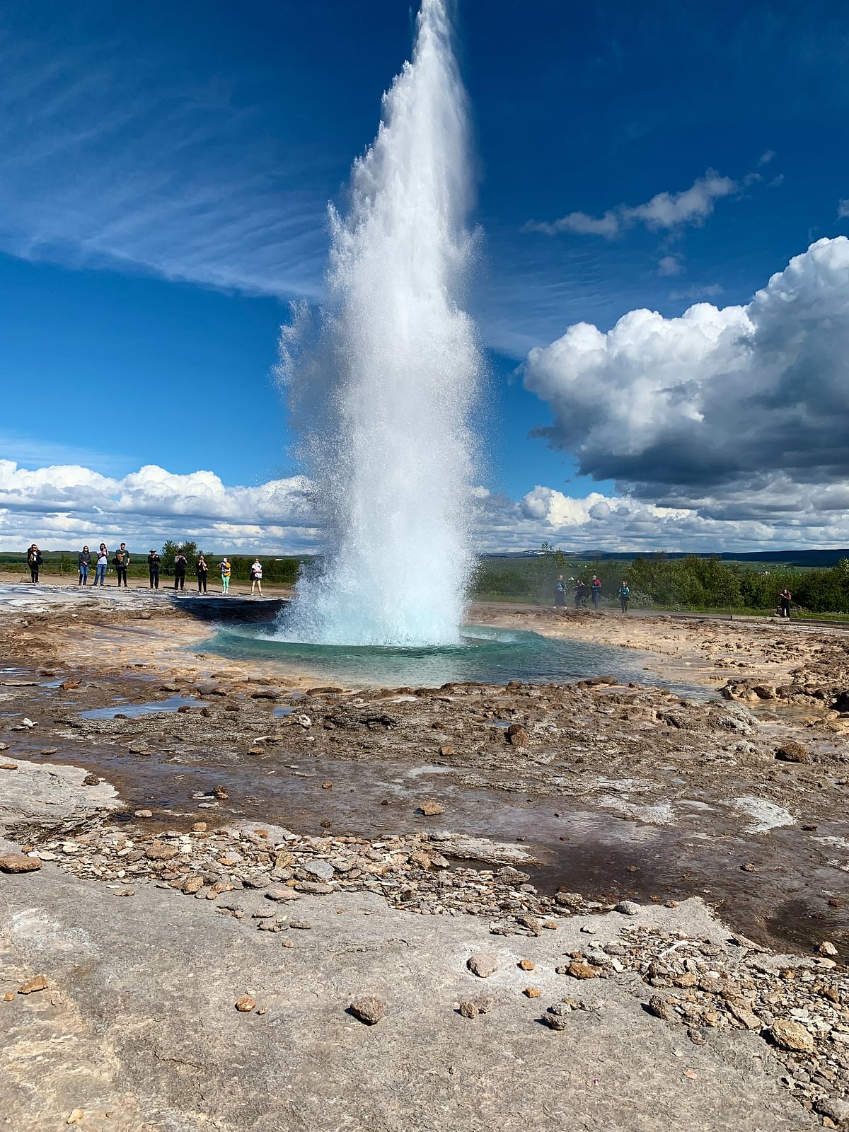 Geyser erupting against a clear blue sky with scattered clouds, surrounded by people watching the natural spectacle.