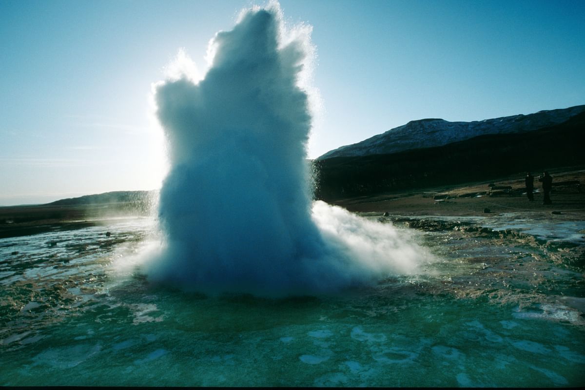 Erupting geyser in Iceland releasing steam and water into the air against a clear blue sky, surrounded by landscape.