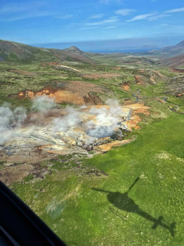 Helicopter shadow over geothermal landscape with steam, vibrant green fields, and hills under a clear blue sky.