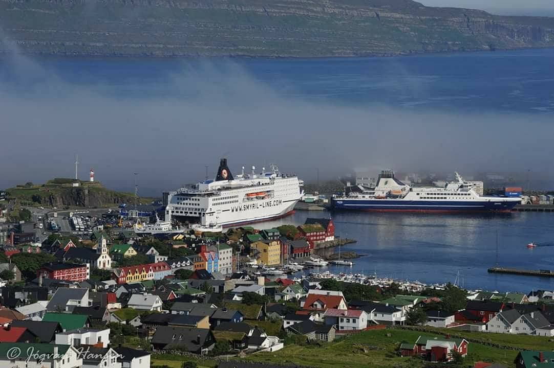 Coastal town with colorful houses and two ferries docked in a scenic harbor, mountains in the background.