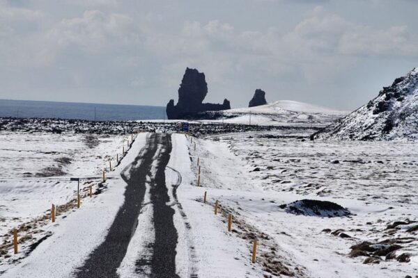 Snow-covered road leading to jagged rock formations by the sea under a cloudy sky.