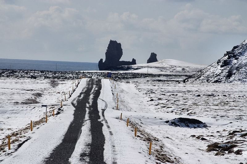 Snow-covered road leading to jagged rock formations by the sea under a cloudy sky.