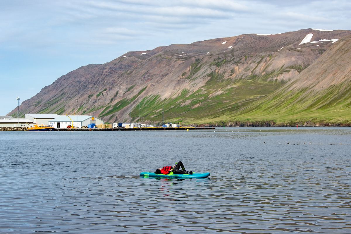 Kayaker paddling on calm waters near a mountain landscape and harbor buildings.