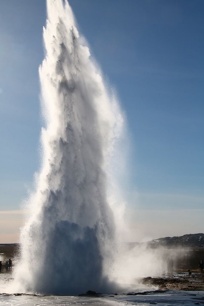 Geyser erupting with powerful water jet against clear blue sky, surrounded by rocky terrain and distant hills.