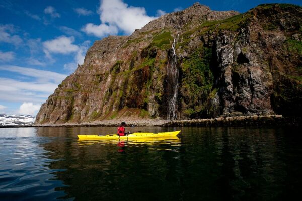 Person kayaking near a rocky cliff with waterfall, under a blue sky.