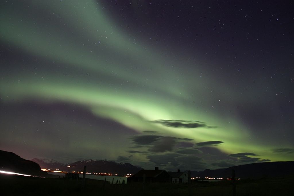 Northern Lights illuminate the night sky over a lakeside village with distant mountains and scattered stars.