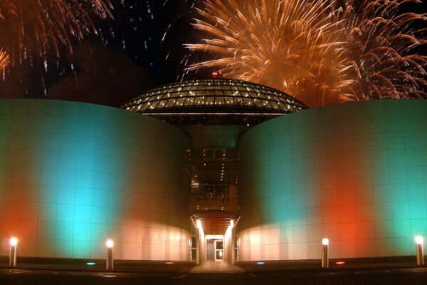 Modern building with a glass dome, illuminated in green, white, and red, under a night sky with vibrant fireworks.