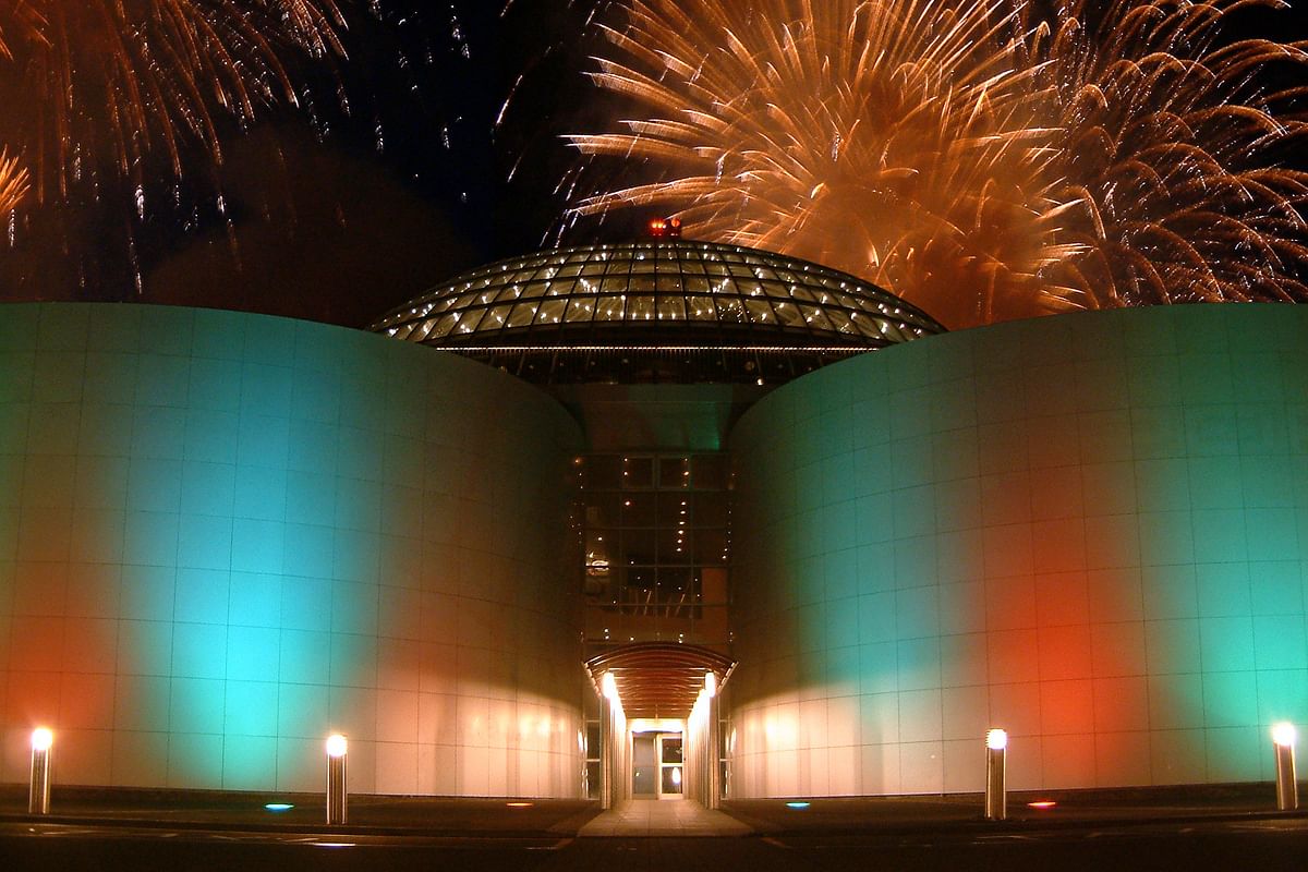 Modern building with a glass dome, illuminated in green, white, and red, under a night sky with vibrant fireworks.