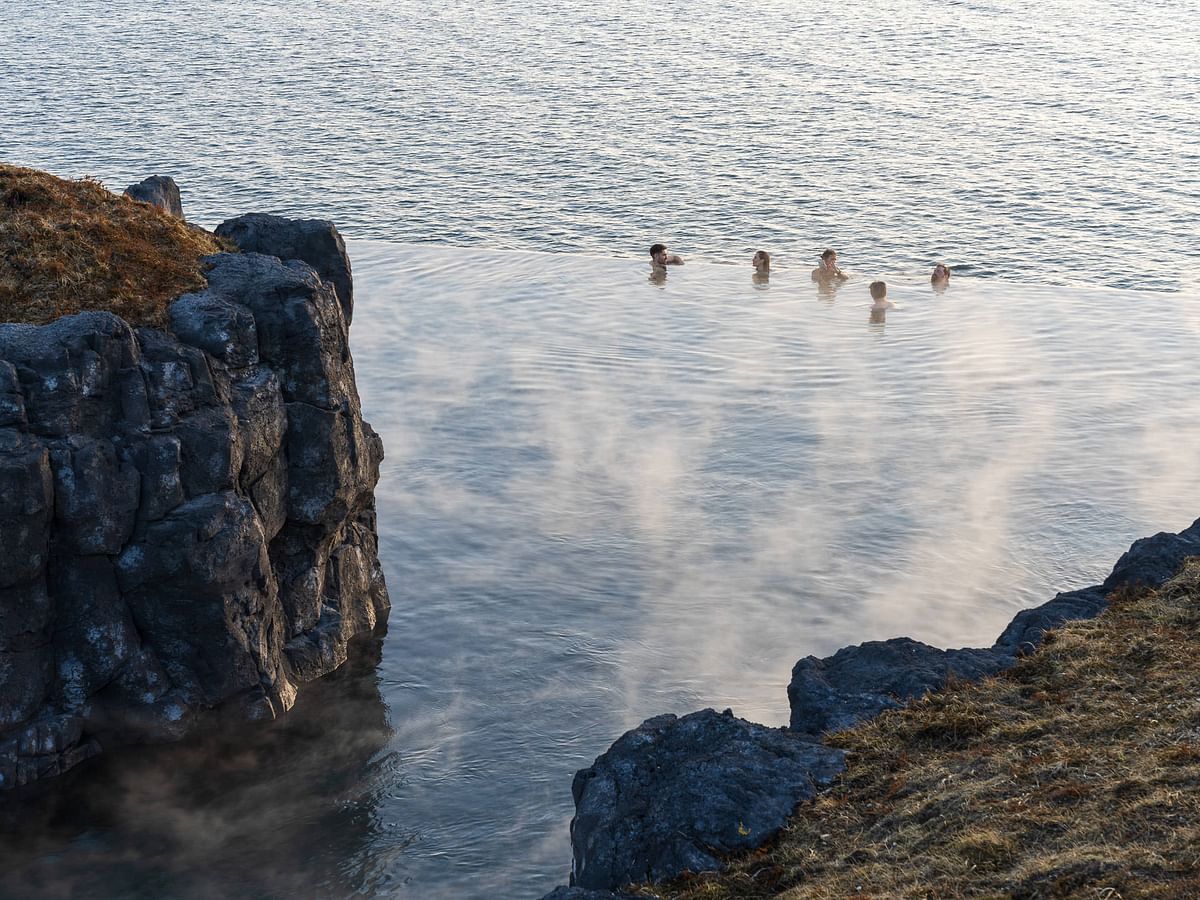 People enjoying a swim in a natural volcanic hot spring beside rocky cliffs with steam rising from the water.