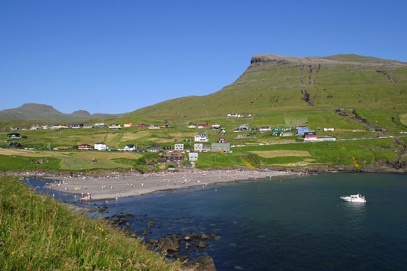 Sunny beach scene with green hills, village houses, and a boat in a scenic coastal landscape under blue sky.