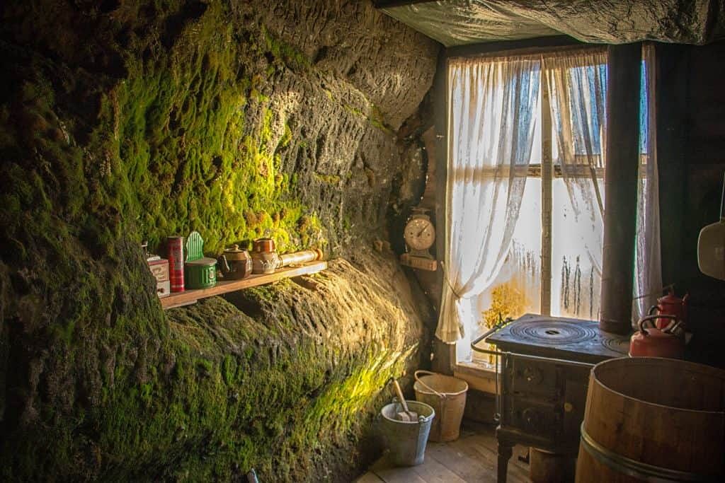 Rustic stone-walled kitchen with moss, antique stove, and window with lace curtains. Cozy vintage interior scene.