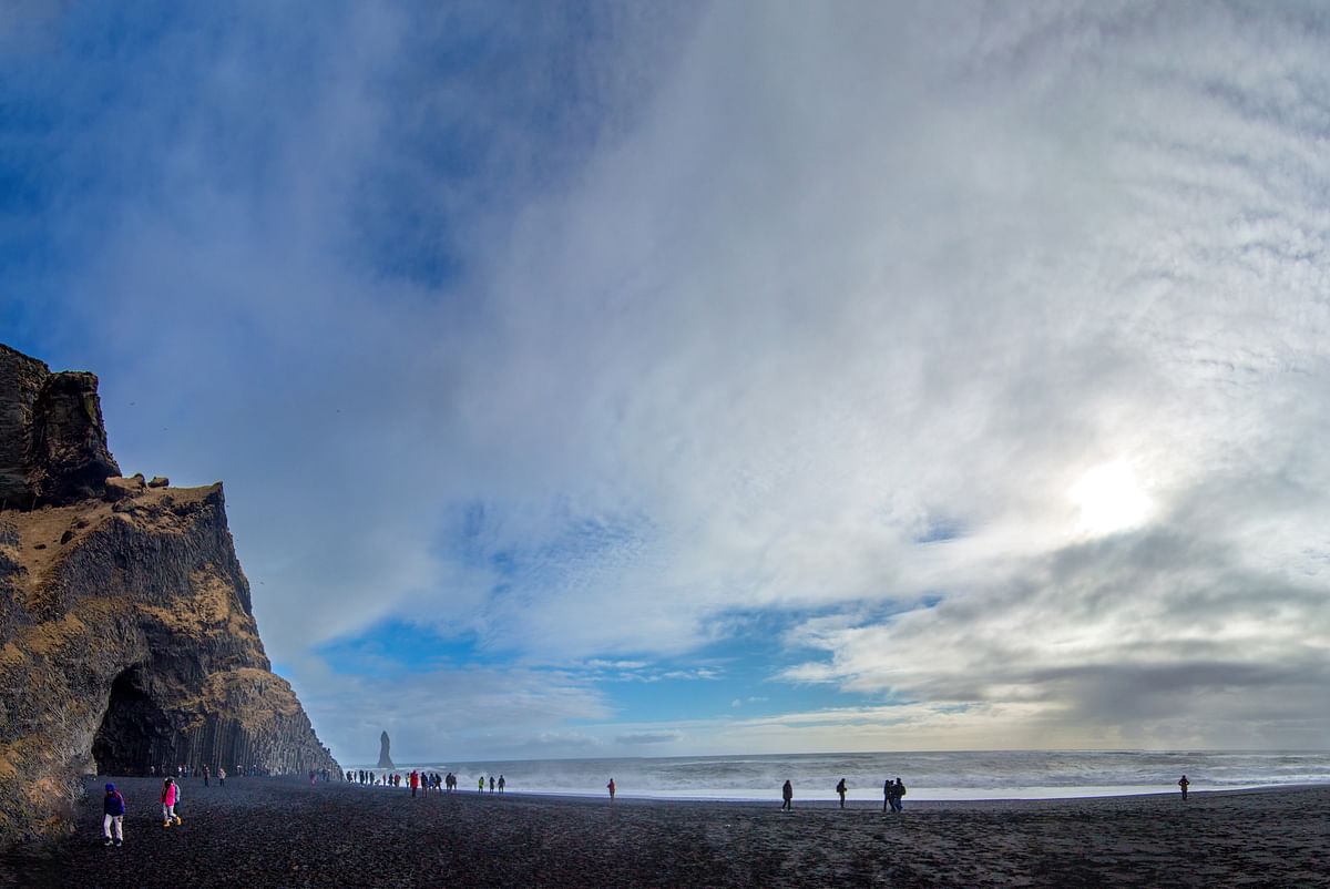 People walking on a black sand beach with steep cliffs under a cloudy sky, ocean waves in the background.