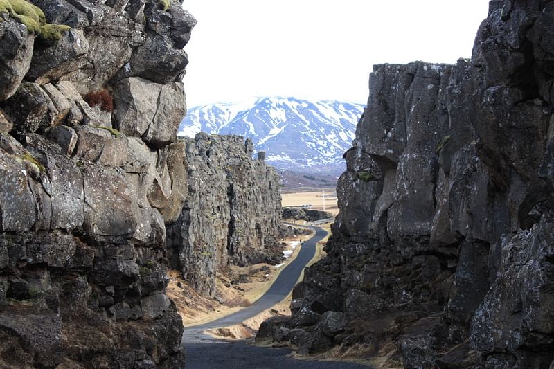 Scenic view of rugged cliffs and a winding road leading to a snow-capped mountain under a clear sky.