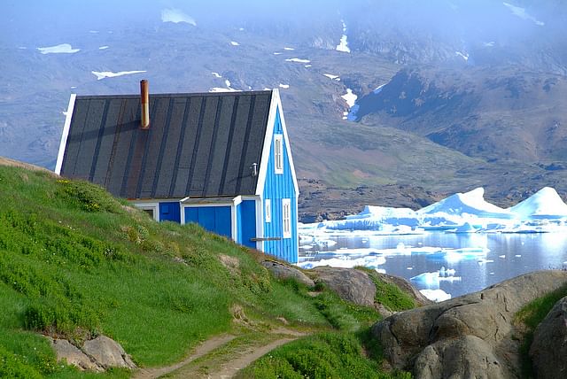 Blue house by a serene lake with icebergs and mountain backdrop, under a clear sky in a tranquil landscape.