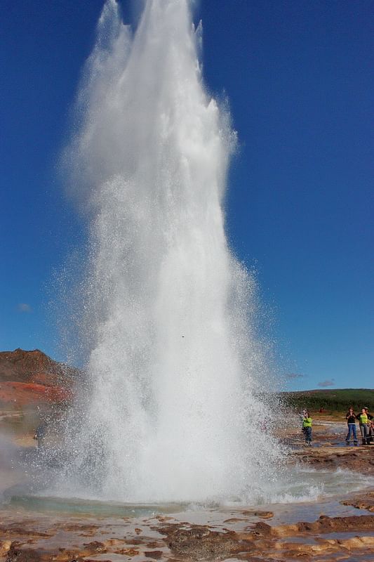 Powerful geyser erupting under clear blue sky, with onlookers capturing the natural spectacle.