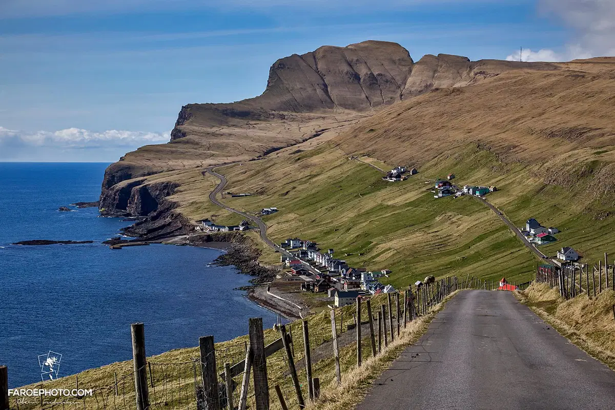 Scenic view of a coastal village in the Faroe Islands with green hills and cliffs by the ocean under a blue sky.