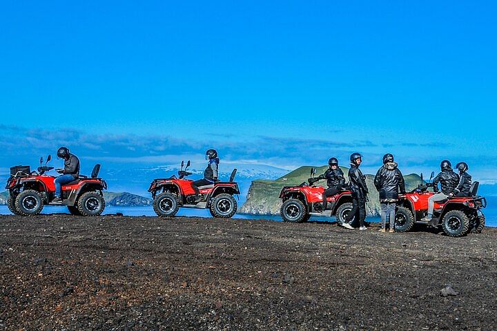 People riding red ATVs on a scenic coastal terrain under a clear blue sky.