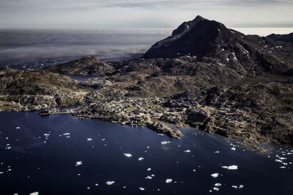 Aerial view of a coastal village with mountain backdrop, Greenland fjord, and scattered icebergs on a clear day.