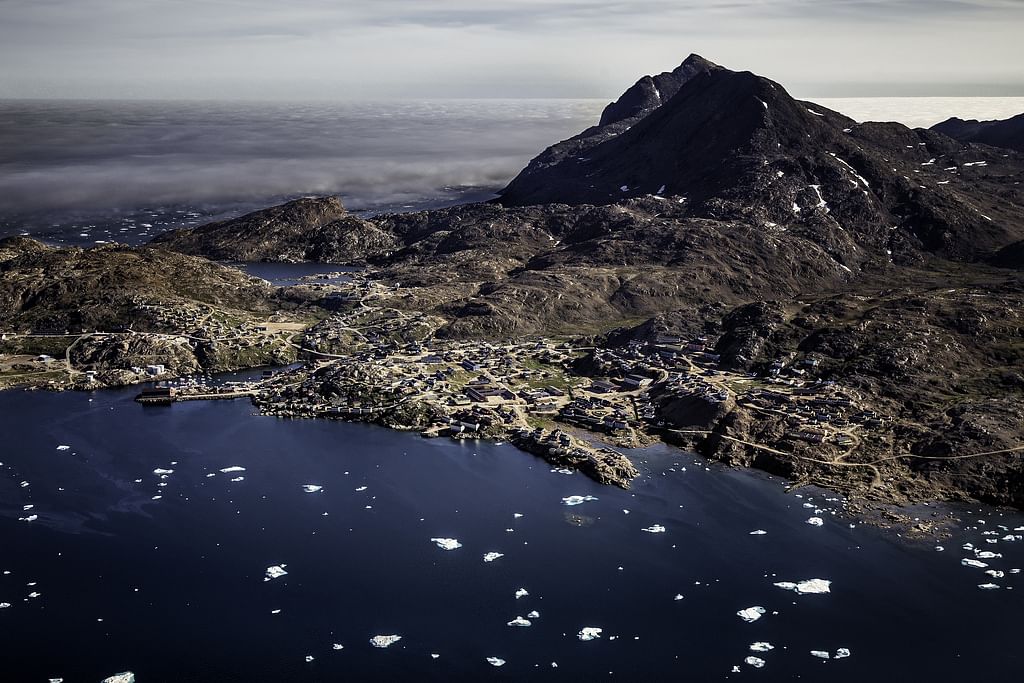 Aerial view of a coastal village with mountain backdrop, Greenland fjord, and scattered icebergs on a clear day.