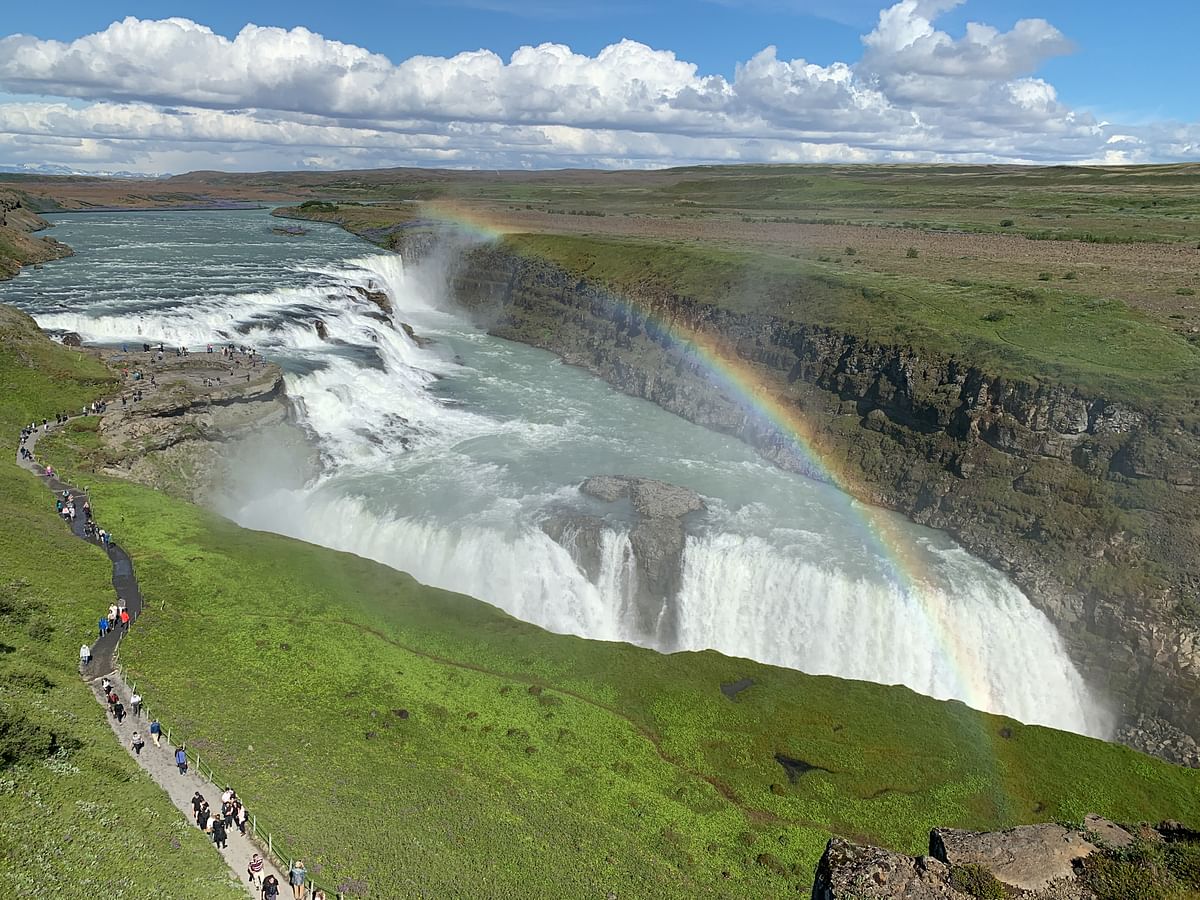 Majestic waterfall with a vibrant rainbow arching over green landscape and tourist path on a sunny day.