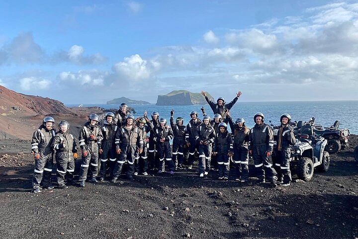 Group of people in protective gear posing on a rocky coast with ATVs, ocean, and islands in the background.