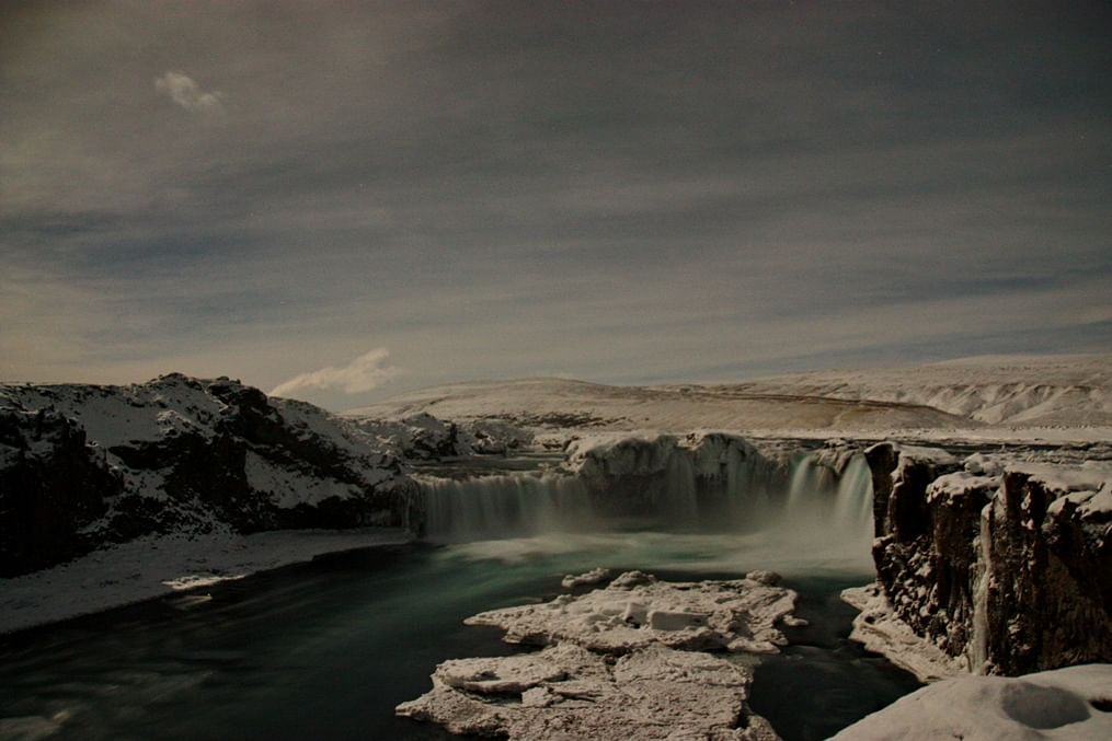 Snow-covered landscape with a waterfall cascading into a river under a cloudy sky. Peaceful and serene winter scene.
